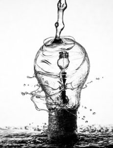 water flowing down onto a lightbulb whose inner filaments are liquifying from the change and throwing off droplets, black and white