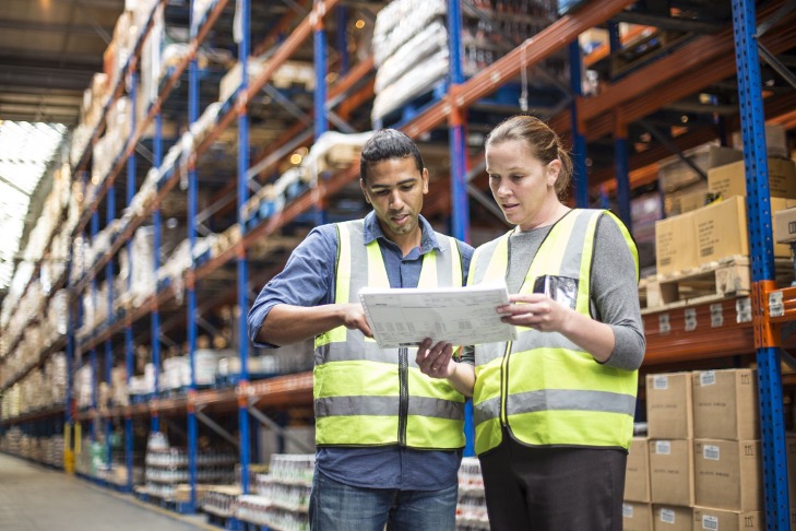 Two workers in a warehouse wearing safety vests looking at a paper report.
